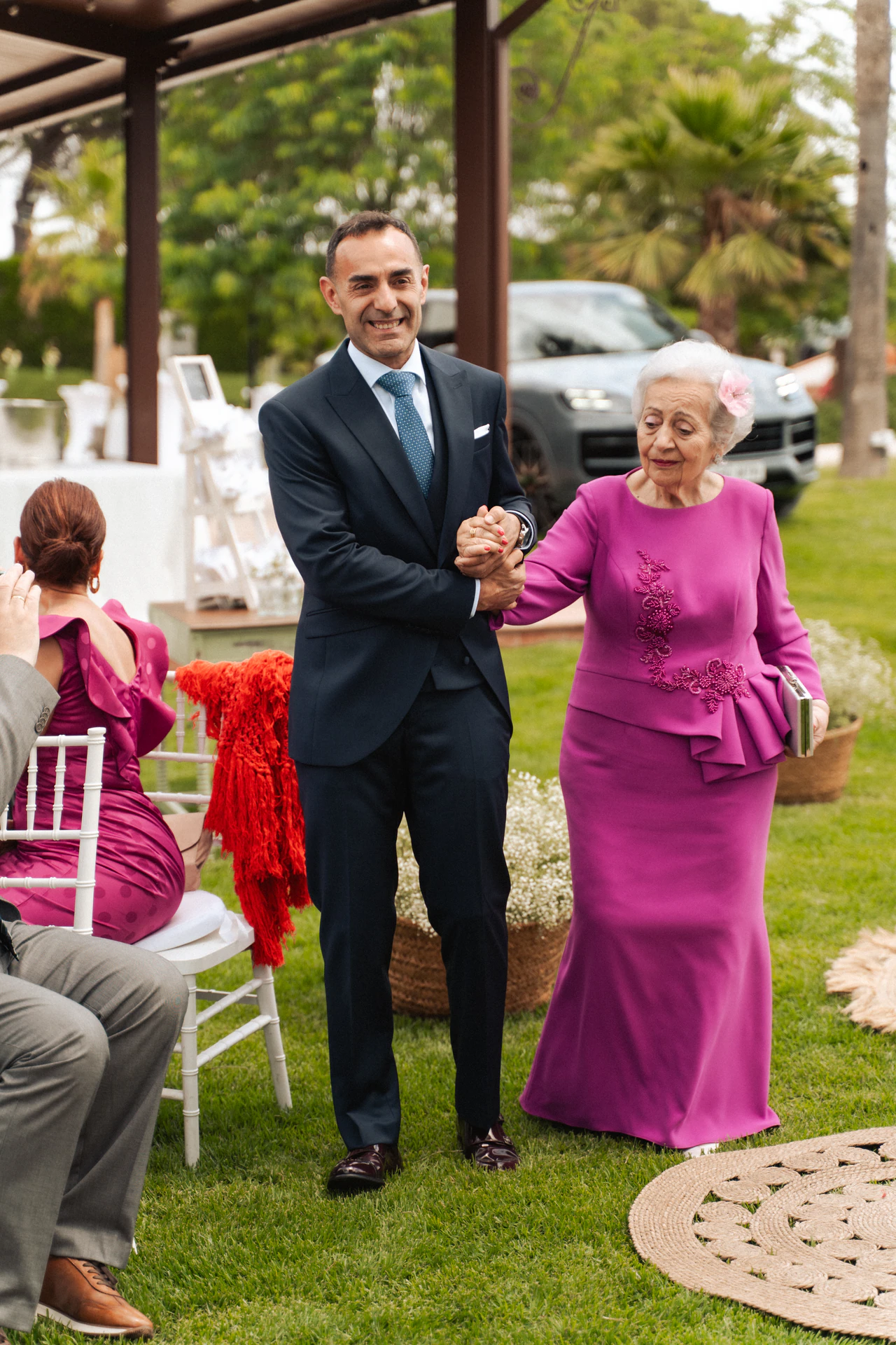 Fernando llega a la ceremonia en el jardín del brazo de una mujer mayor vestida de fucsia