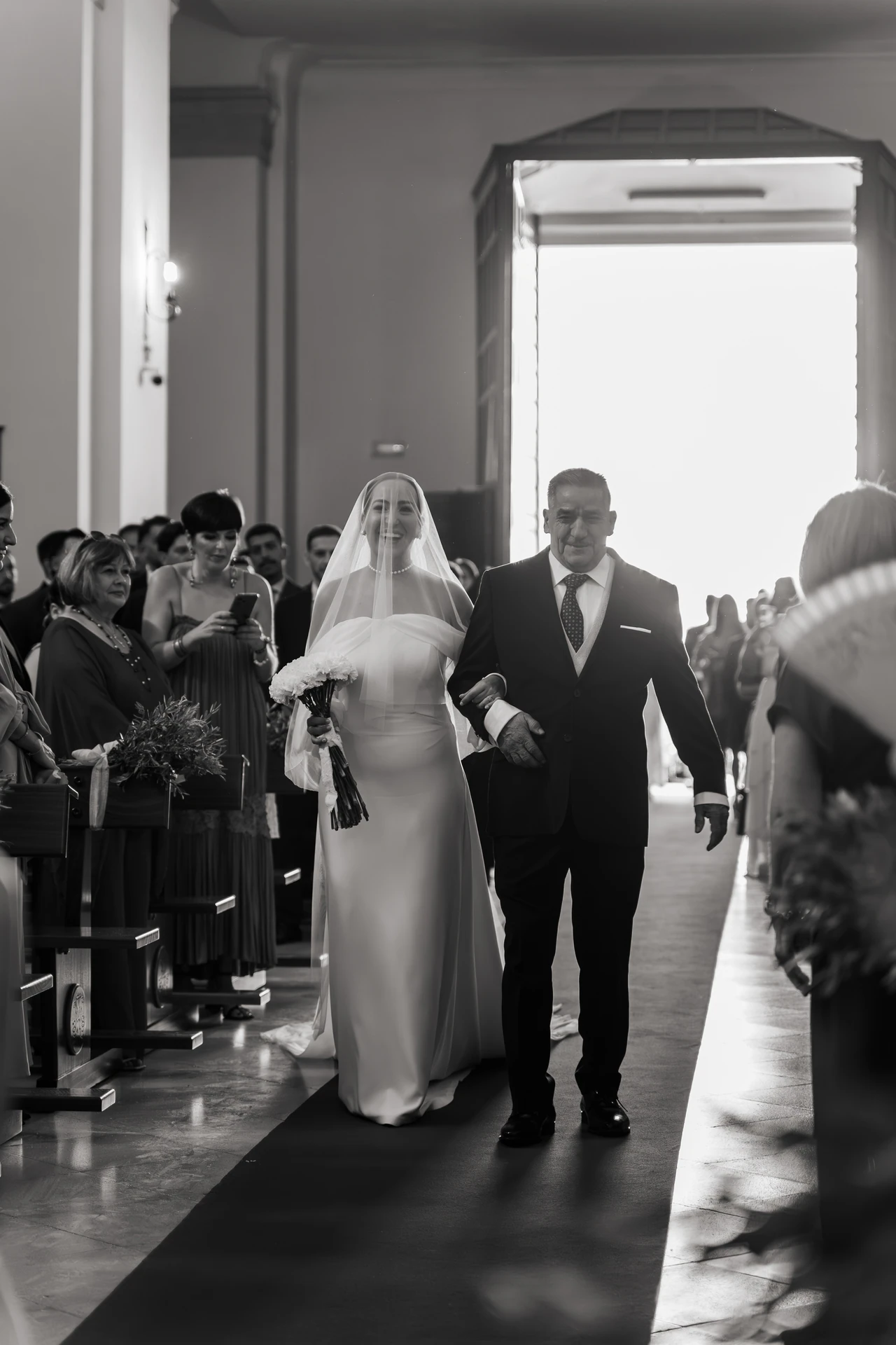María entra en la iglesia del brazo de un hombre, en blanco y negro