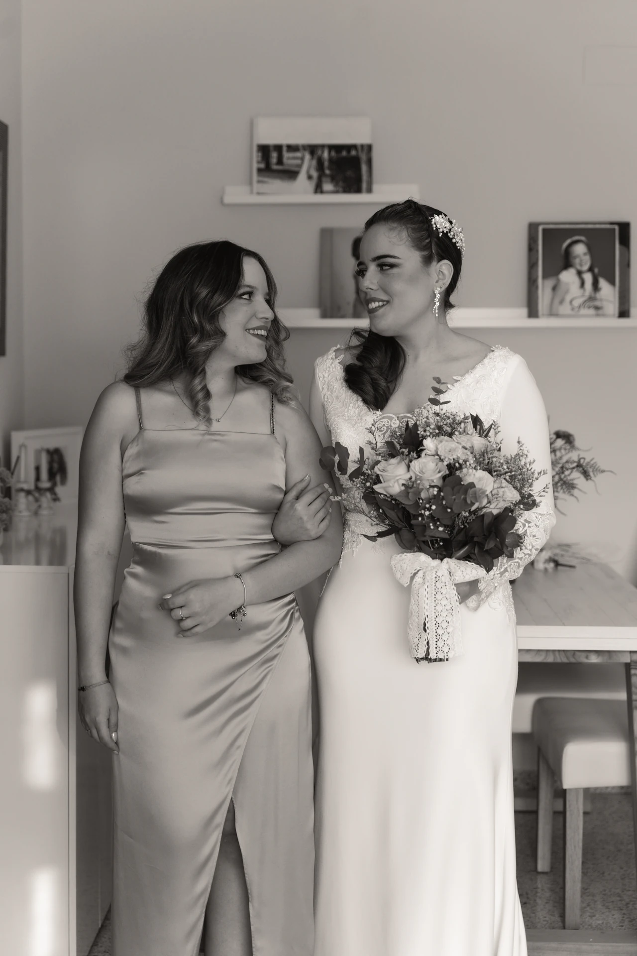 Una de las novias, con su ramo, junto a una amiga de vestido satinado, en blanco y negro