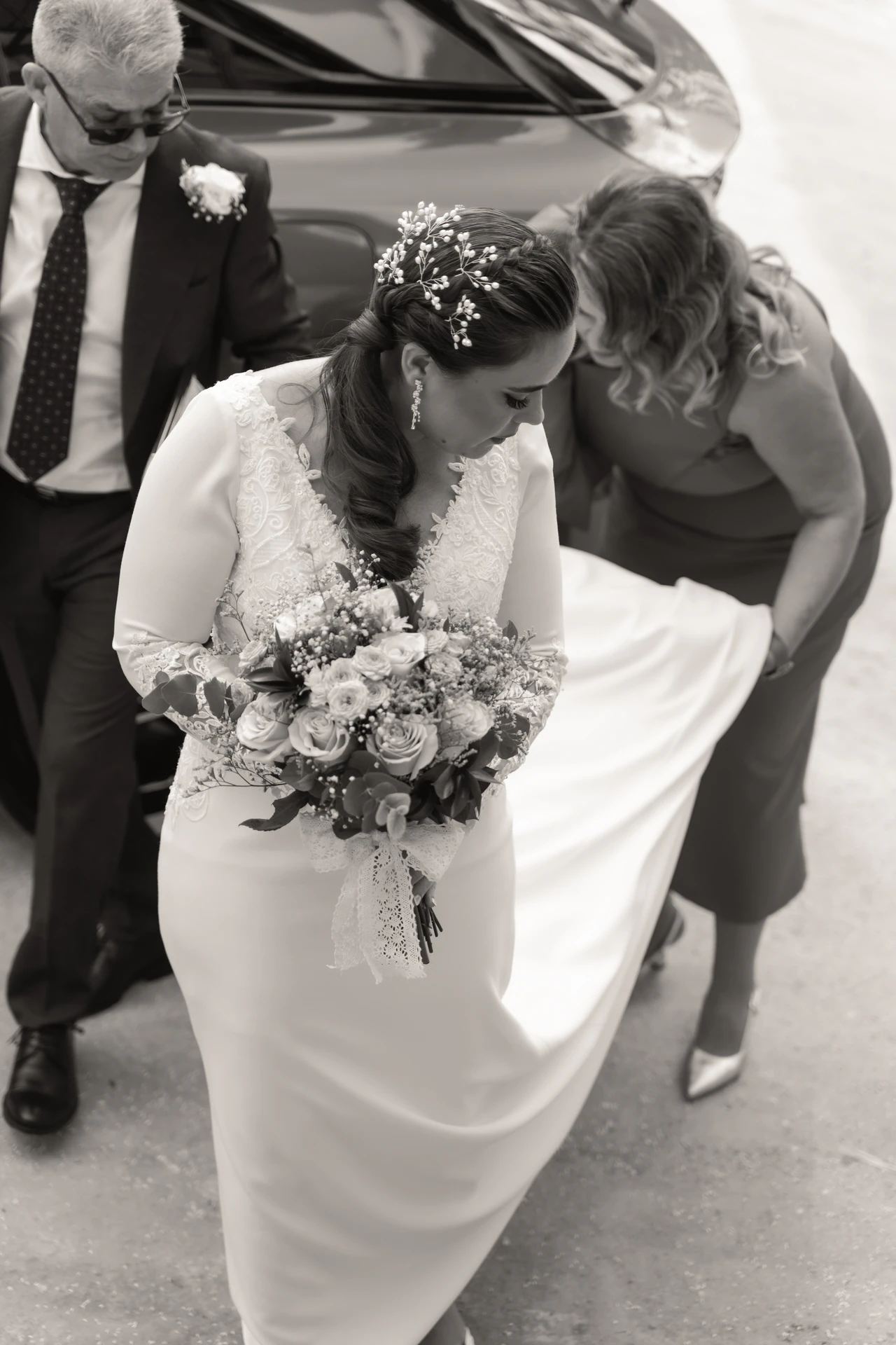 Ayudan a una de las novias con el vestido al bajar del coche, en blanco y negro